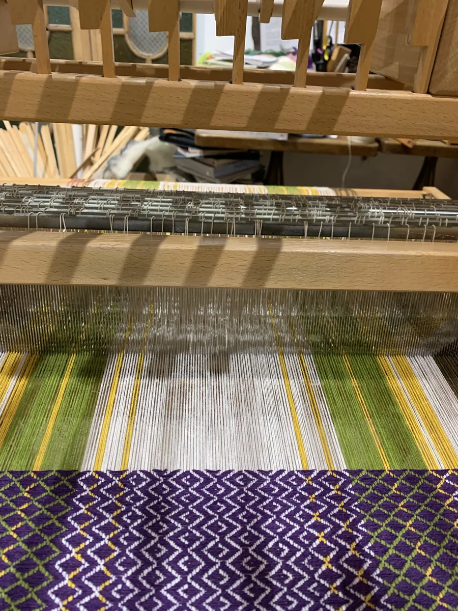 Gros plan d'un tissage en cours sur un métier à tisser à bras : chaîne écrue, motifs violets, verts et jaunes