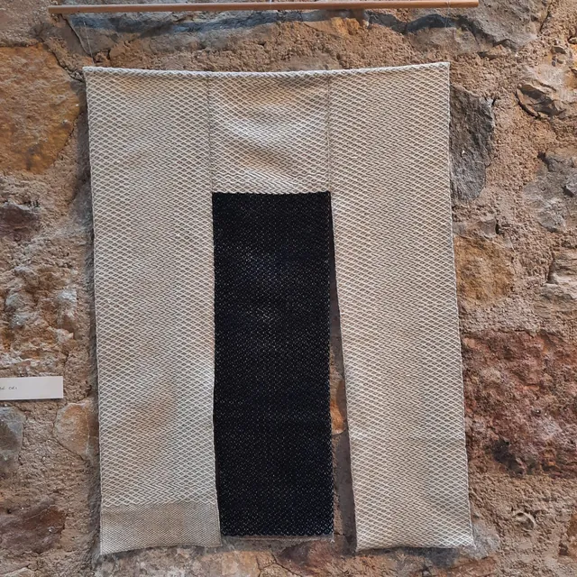 Tenture tissée main écrue et noire, accrochée sur un mur de pierre